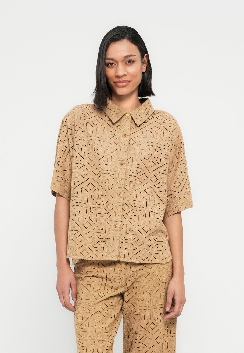 Chemise à manches courtes couleur sable avec un motif géométrique découpé, col à boutons et fermeture à boutons sur le devant. Tissu texturé avec une coupe décontractée.