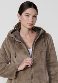 Puhast rjav zip-up kapuca s puhaste teksturo, z vrvico in kontrastno rjavo notranjo podlogo. Ima sproščen kroj in kapuco.