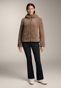 Brun fuskpälsjacka med huva, framdragkedja och vida ärmar, i kombination med svarta utsvängda byxor och vita sneakers.