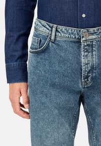 Šviesiai mėlyni džinsai su nudėvėtu paviršiu, klasikinio penkių kišenių dizaino ir mygtukų uždarymu. Matoma džinsinio audinio tekstūra.