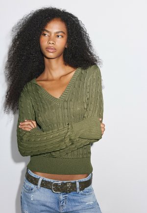 Ung kvinde med krøllet hår iført en grøn kabelstrikket sweater og lyseblå revnede jeans, stående med armene krydset mod en hvid baggrund.