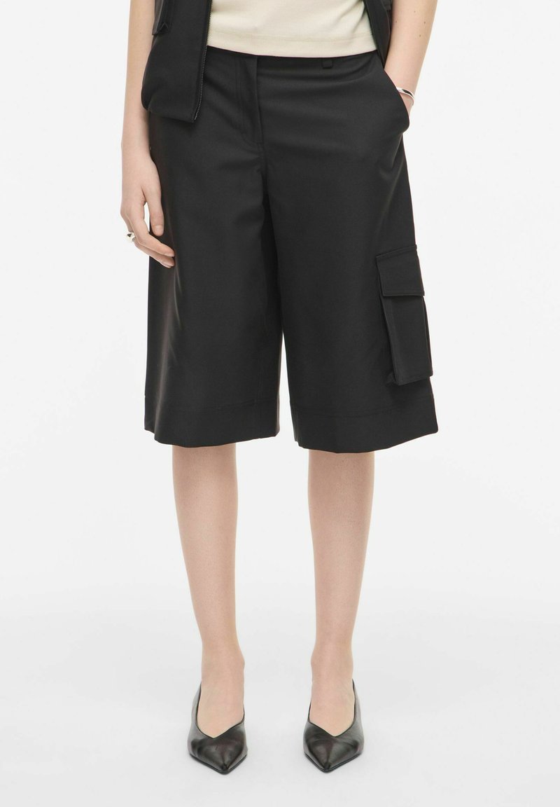 Femme portant un short cargo noir longueur genou avec une poche latérale et des talons noirs pointus, debout les mains dans les poches.