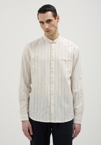 Uomo che indossa una camicia button-up a righe verticali color crema con colletto alla chinesina e pantaloni neri, in piedi su uno sfondo neutro.