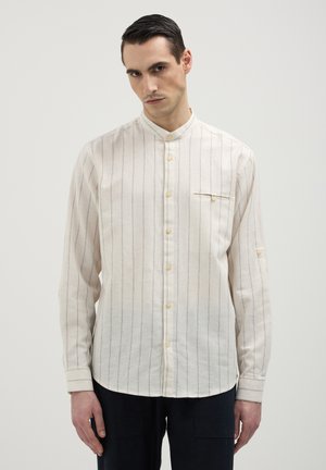 Uomo che indossa una camicia button-up a righe verticali color crema con colletto alla chinesina e pantaloni neri, in piedi su uno sfondo neutro.