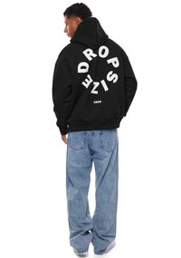 Svart hettegensere med et stort hvitt grafikk som leser "DROP" og "CREW" på baksiden, kombinert med løstsittende lyseblå jeans.