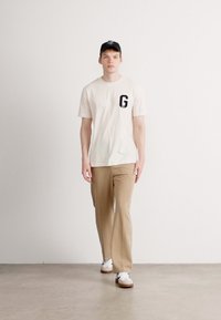 Jeune homme marchant à l'intérieur, portant un pantalon beige, un t-shirt blanc avec un "G" noir sur la poitrine, une casquette noire et des baskets blanches avec des semelles brunes.