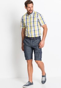 Camicia a maniche corte con colletto a quadri gialli, blu e bianchi, abbinata a pantaloni cargo blu scuro, completata da sneakers navy.