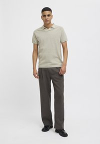 Beige polo shirt med strukturert stoff, kortermede ermer og tre-knappers placket, kombinert med brune pressebukser og svarte sko.