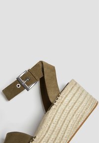 Sandale à talon compensé en suède vert olive avec un bout rond, une bride de cheville avec une boucle en métal, et une semelle en jute tissée.
