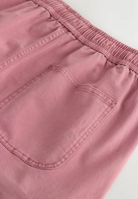 Pantalon en tissu rose avec taille élastique et une poche arrière rectangulaire cousue sur un fond blanc.