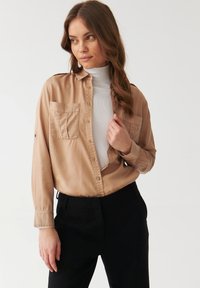 Beige knopenblik shirt van gladde stof, met twee borstzakken en opgerolde mouwen, gedragen over een witte coltrui.