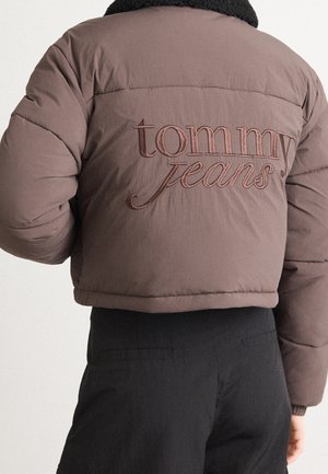 Doudoune marron avec col noir brodé "tommy jeans" dans le dos, portée avec un pantalon noir, vue de dos.