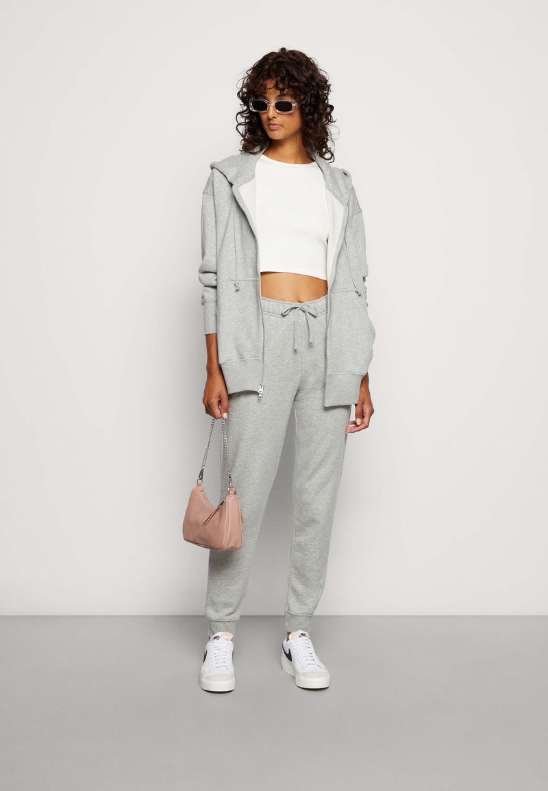 Haut à capuche gris avec cordons de serrage ajustables, associé à un pantalon de survêtement assorti. Le mannequin tient un petit sac à main rose, portant des chaussures blanches et des lunettes de soleil.