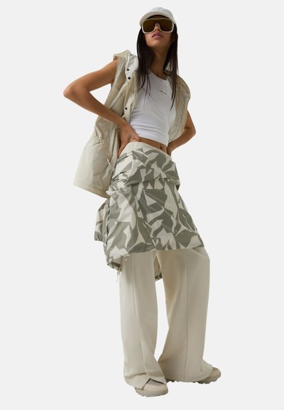 Leichtes, weißes ärmelloses Top, kombiniert mit einer beigen Weste. Der Rock hat ein grün-weißes geometrisches Muster und wird mit weiten cremefarbenen Hosen getragen.