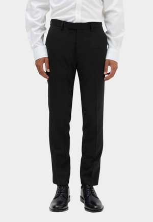 Man wearing black formal trousers, black dress shoes, and a white buttoned shirt standing against a plain background.