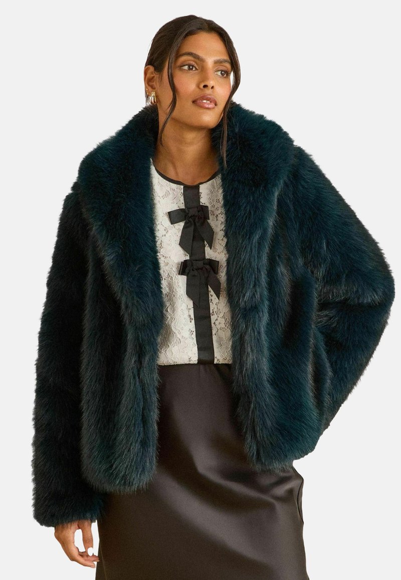 Tūtiņu teal mākslīgās kažokādas jaka ar plašu apkakli, maigu tekstūru un brīvu piegriezumu, uzvilkta virs baltas mežģīņu blūzes ar melnā svītru akcentiem.