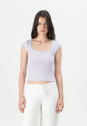 Jeune femme aux cheveux noirs longs portant un haut lilas à manches courtes côtelé et un pantalon blanc à cordon de serrage devant un fond blanc uni.