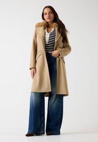 Cappotto di lana beige con un colletto in pelliccia sintetica, abbinato a un top a righe bianco e nero e a jeans blu a gamba larga. Tessuto testurizzato e design strutturato.
