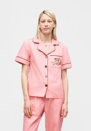 Moteris vilki šviesiai rožinį trumpomis rankovėmis miego drabužių komplektą su juodais apvadais ir gėlių siuvinėjimu ant krūtinės kišenės, stovinti prie paprasto fono.