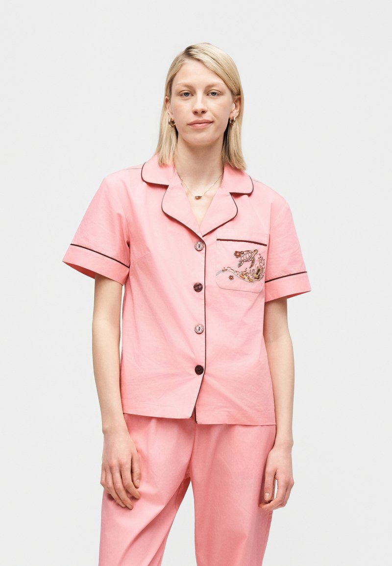 Frau trägt einen hellrosa Pyjama mit kurzen Ärmeln, schwarzer Paspelierung und floraler Stickerei auf der Brusttasche, sie steht vor einem einfarbigen Hintergrund.