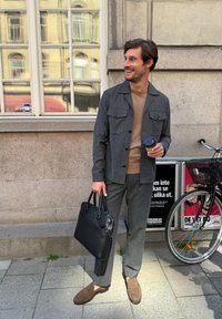 Grijs gestreepte shirtjack over een tan kleurige trui, gecombineerd met grijze broek en bruine suède instappers. Zwart leren tas en koffiekopje in de hand.
