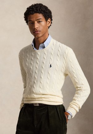 Jeune homme aux cheveux bouclés portant un pull torsadé crème sur une chemise bleu clair, un pantalon sombre et une montre-bracelet, debout avec une main dans la poche.