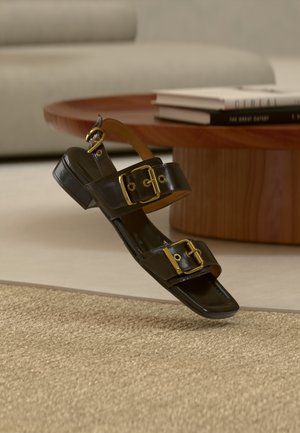 Μαύρο δερμάτινο σανδάλι με δύο χρυσά αγκράφες και χαμηλό τακούνι, τοποθετημένο μπροστά από ένα στρογγυλό ξύλινο τραπεζάκι με στοίβες βιβλίων σε εσωτερικό χώρο.