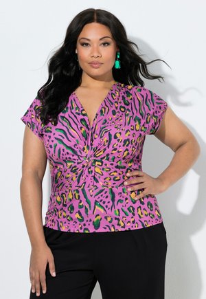 Frau mit langen dunklen Haaren, die grüne Ohrringe trägt und ein rosafarbenes, gemustertes Kurzarmoberteil mit Twist-Detail, posiert mit einer Hand in der Hüfte.