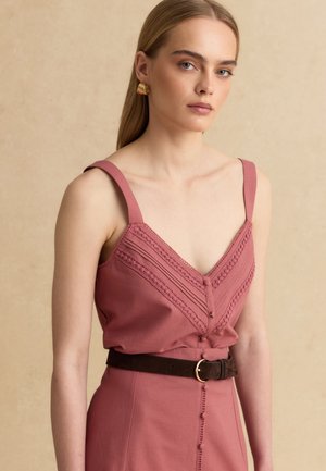 Giovane donna che indossa un abito senza maniche rosa polvere con dettagli ricamati e una cintura scura, posando su uno sfondo beige.