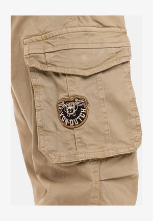Poche de pantalon cargo kaki avec rabat et patch logo crâne Von Dutch brodé en fils noir, blanc et marron.