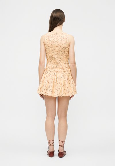 Γυναίκα με μακριά καστανά μαλλιά, φοράει αμάνικο, ροδακινί φόρεμα με μικρές κόκκινες τελείες και καφέ παπούτσια με κορδόνια, στέκεται πλάτη σε λευκό φόντο.