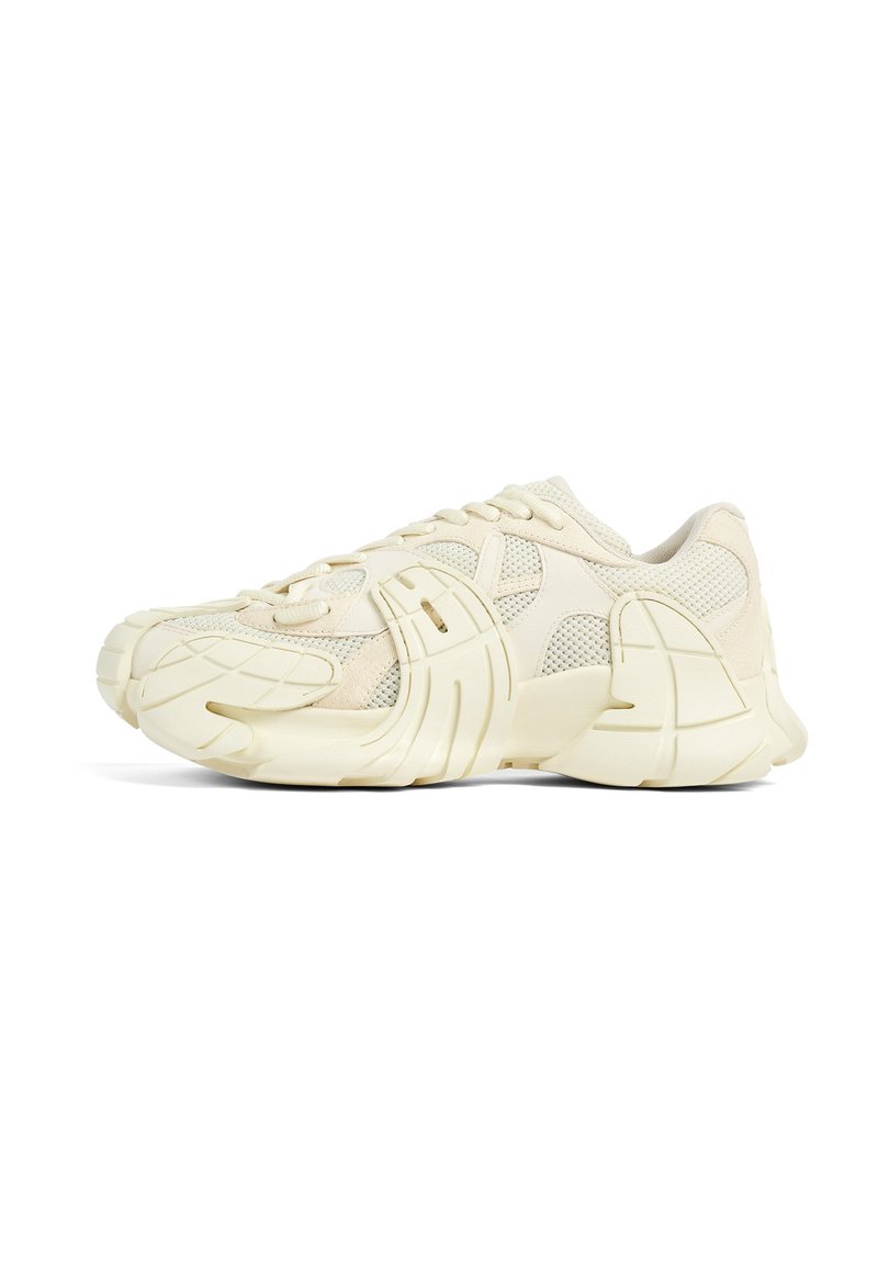 Cream-colored sneaker with a textured mesh upper, prominent rubber sole, and unique sculpted shape. Features lacing system and padding.