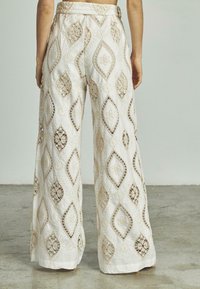 Pantaloni bianchi a gamba larga con elaborati ricami beige in un motivo a diamante, con dettagli in pizzo visibili e una texture di tessuto liscia.