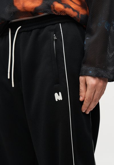 Gros plan sur une personne portant un pantalon de survêtement noir avec des cordons blancs, une poche zippée, une bande blanche et un "N" blanc brodé sur la cuisse.