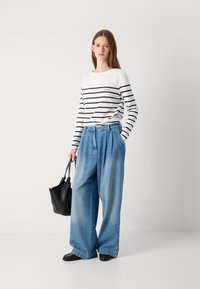 Gestreepte witte en marineblauwe blouse met lange mouwen en een rood logo, gecombineerd met lichtblauwe wijde jeans en een zwarte tote bag. Zwarte schoenen maken de look compleet.
