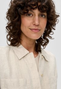 Light beige linen shirt with a collar, two front pockets, and button closure. Soft texture and natural fiber appearance.