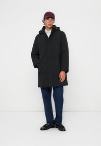 Zwarte parka-jack met capuchon, met een rechte snit en voorzakken, gedragen over een licht shirt en blauwe jeans. Stof lijkt glad.
