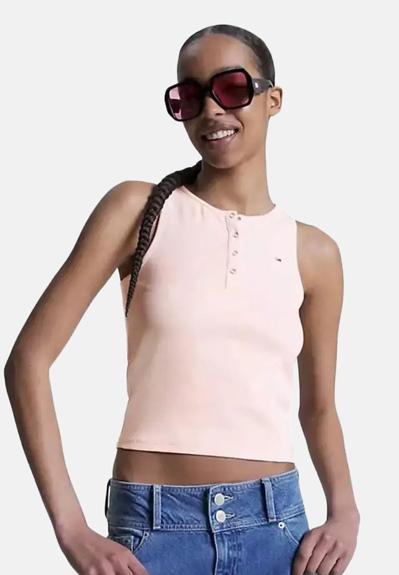 Femme aux cheveux tressés portant de grandes lunettes de soleil carrées, un crop top rose clair sans manches et un jean taille haute bleu, souriante.