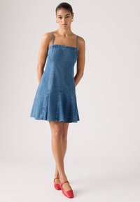 Γυναίκα στέκεται φορώντας αμάνικο μπλε φόρεμα τζιν με φαρδιά φούστα και κόκκινα παπούτσια Mary Jane σε λευκό φόντο.