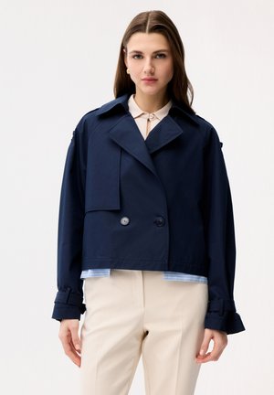 Młoda kobieta ubrana w krótką granatową kurtkę dwurzędową z pagonami, beżowe spodnie oraz białą koszulę z kołnierzykiem, stojąca na białym tle.