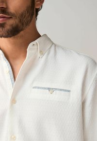Chemise blanche à motif avec une finition texturée, présentant un col boutonné et une poche poitrine rehaussée de bleu avec un détail de bouton.