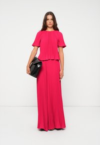 Fuchsia-gefalteter Jumpsuit mit kurzen Ärmeln, der ein geschichtetes Design aufweist, kombiniert mit einer schwarzen Handtasche. Glatte Textur und weit geschnittene Beine.
