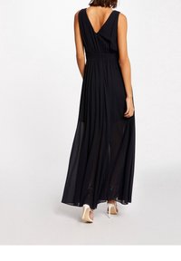 Maxikjol i svart, med djup V-rygg, samlad midja och genomskinligt tyg. Stängs med en knapp i ryggen, kombinerad med vita klackar.