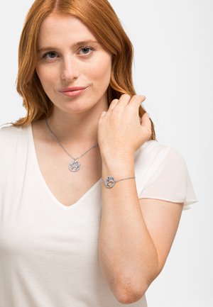 Ensemble de bijoux en argent comprenant un collier avec pendentif et un bracelet ornés de motifs d'arbre circulaires. Porté sur un t-shirt blanc à col en V.