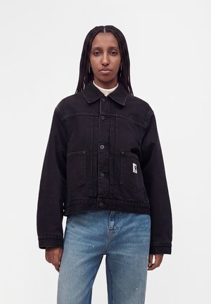 Woman with long braided hair wearing a black buttoned jacket with front pockets and light blue jeans, standing against a plain background.