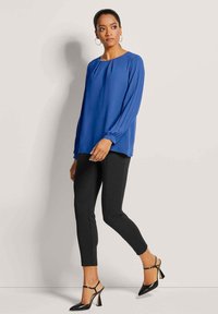 Blaues langärmliges Blusenshirt mit gefalteten Details, kombiniert mit schwarzen, eng anliegenden Hosen und schwarzen Absatzschuhen. Glatter Stoff und maßgeschneiderter Schnitt.