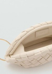 Una borsa a tracolla beige intrecciata con una maniglia metallica circolare, che mostra l'imbottitura interna e un'etichetta discreta.