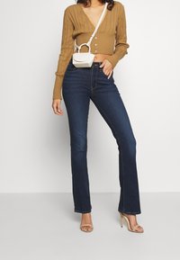 Brun ribbad långärmad topp med V-urringning och knäppning, parat med mörkblå utsvängda jeans och nudefärgade remsandaler. Liten vit handväska.