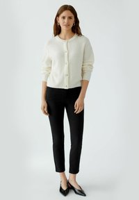 Weißer gestrickter Cardigan mit Rundhalsausschnitt und Knopfverschluss, kombiniert mit schwarzen engen Jeans und schwarzen spitzen Ballerinas.