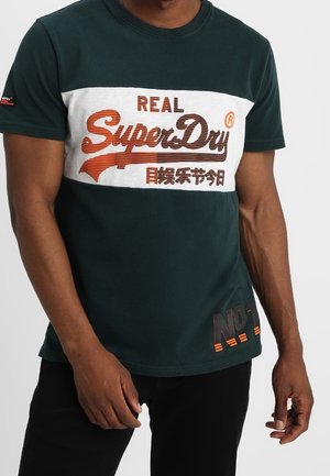 Dark green cotton T-shirt featuring a white panel with "REAL SuperDry" text in orange and brown, textured accents, and "No.1" detail at the hem.